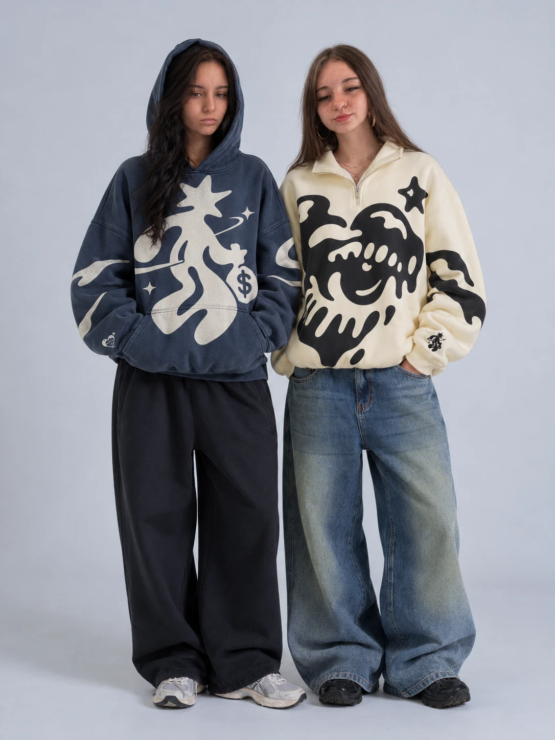 Two people side by side: a washed navy hoodie with the cream creature, and a cream half-zip with the black melting heart.